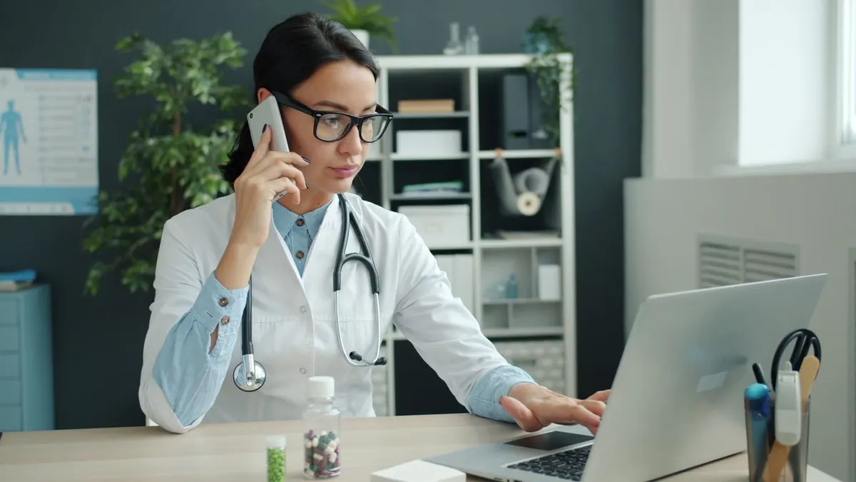 Una médica concentrada realiza varias tareas a la vez con un ordenador portátil y un teléfono en un entorno de oficina