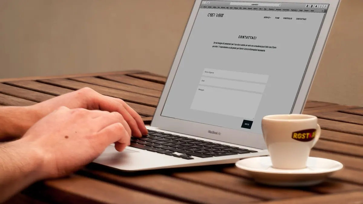 Primer plano de manos escribiendo en un laptop junto a una taza de café en un escritorio de madera.