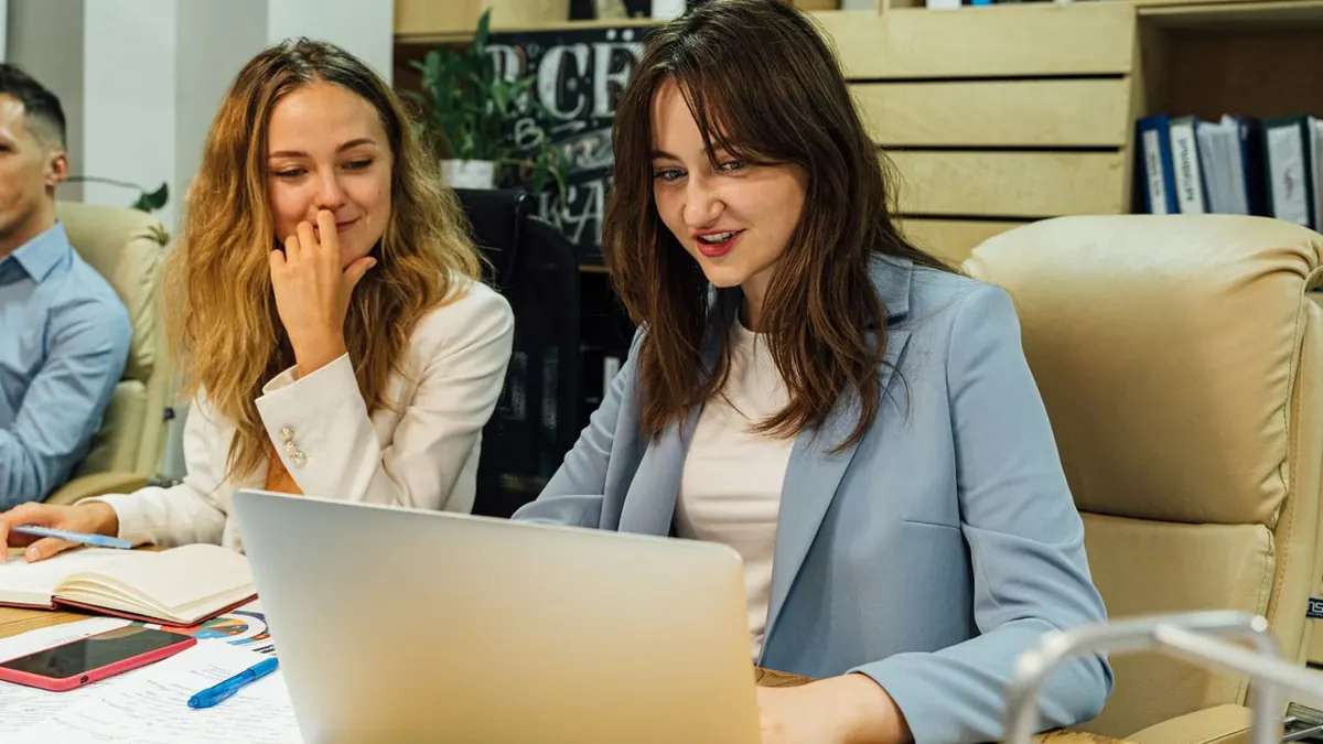 Dos mujeres de negocios con vestimenta corporativa trabajando en un portátil en una oficina moderna.