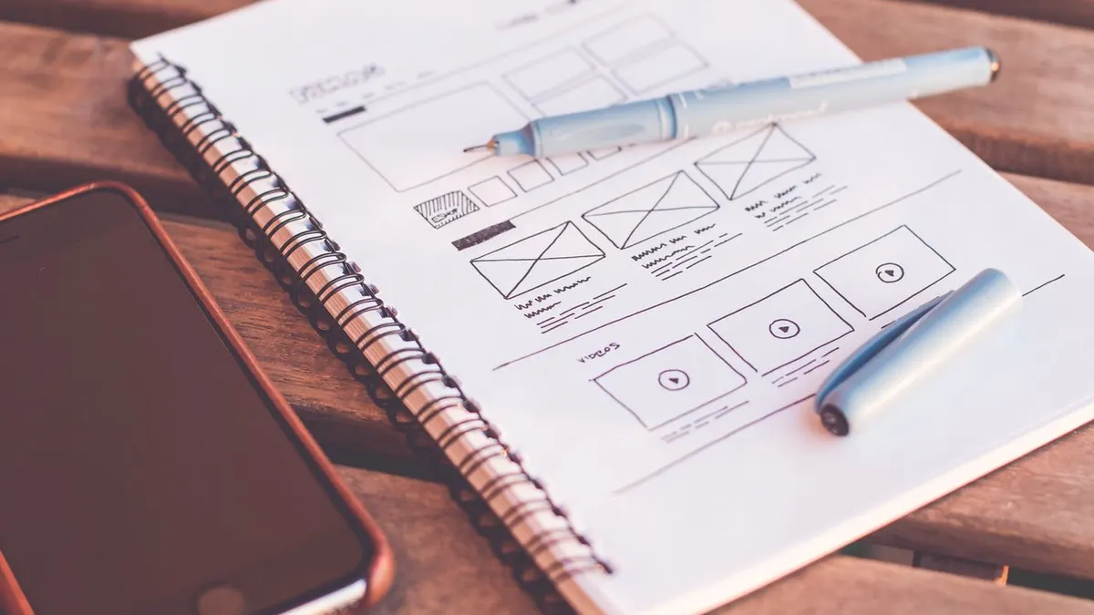 Primer plano de un cuaderno con bocetos en wireframe y un teléfono inteligente sobre un escritorio de madera.
