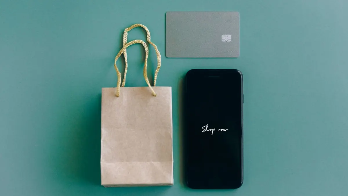 Vista superior de un smartphone con el texto 'Compra ahora', tarjeta de crédito y bolsa de papel sobre fondo verde.